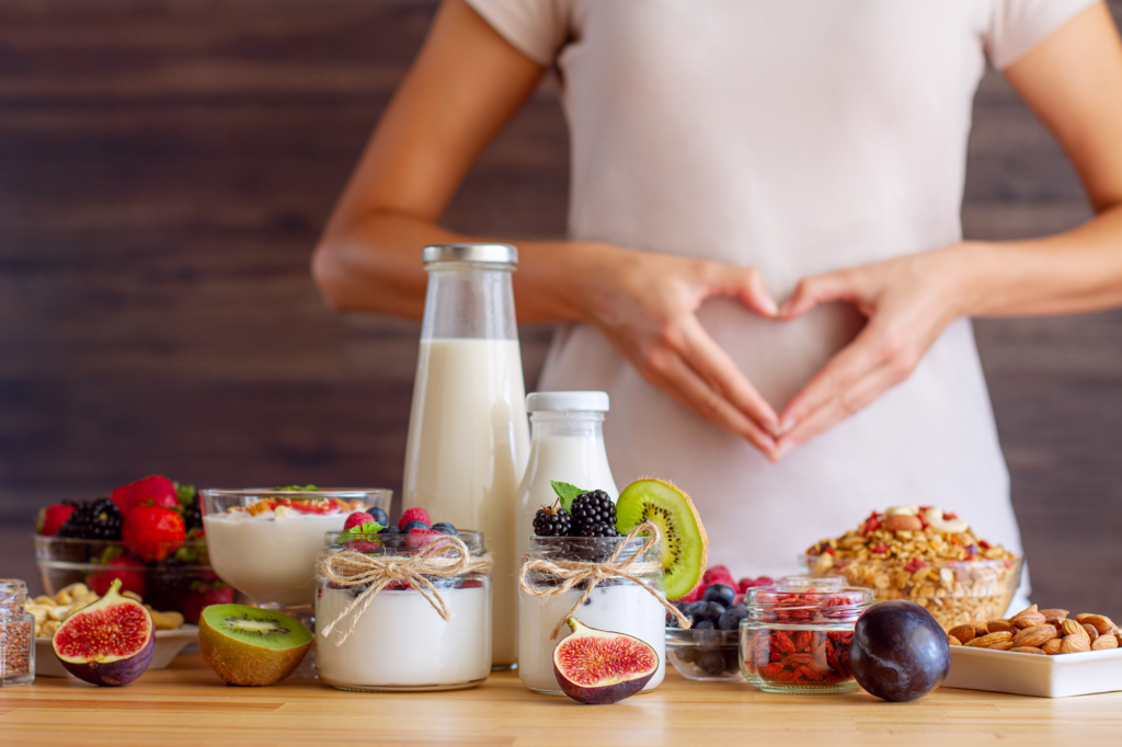 Eine Frau formt ein Herz mit ihren Händen vor ihrem Bauch, im Vordergrund stehen Lebensmittel.