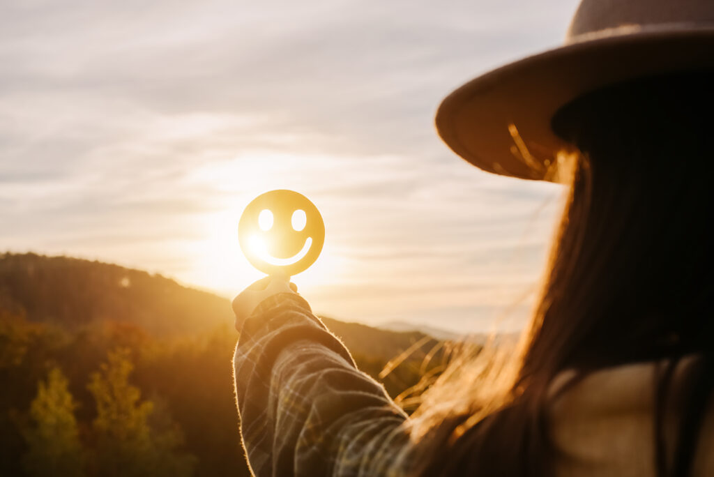 Eine Frau hält einen lächelnden Smiley in den Sonnenaufgang.